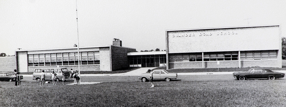 Black and white photograph of Quander Road Elementary School taken in 1969. The building is smaller than Belle View and is mostly one-story tall. Children can be seen playing in front of the school. 