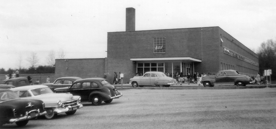 Black and white photograph of Belle View Elementary School taken in 1954 during a fire insurance survey of school properties for the Fairfax County School Board. Students can be seen entering the building and the parking lot is full of late-1940s and early-1950s era cars.